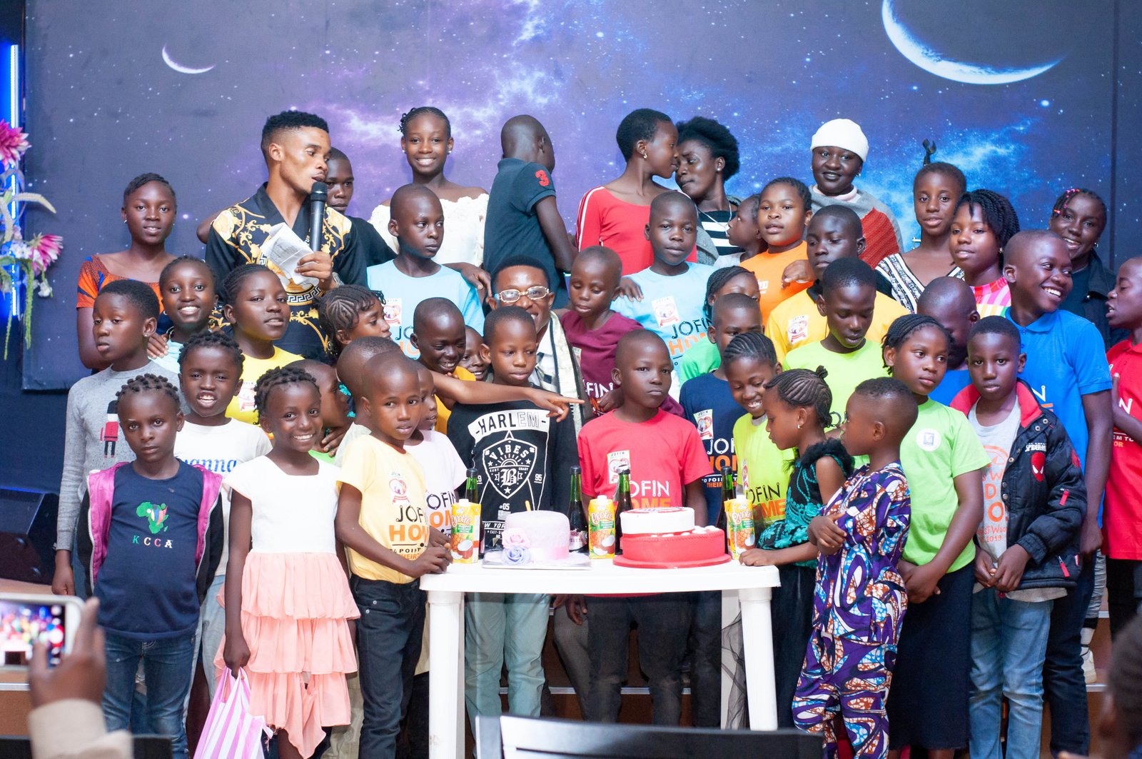 A large group of JOFIN children in colourful T-shirts gathered around celebration cakes