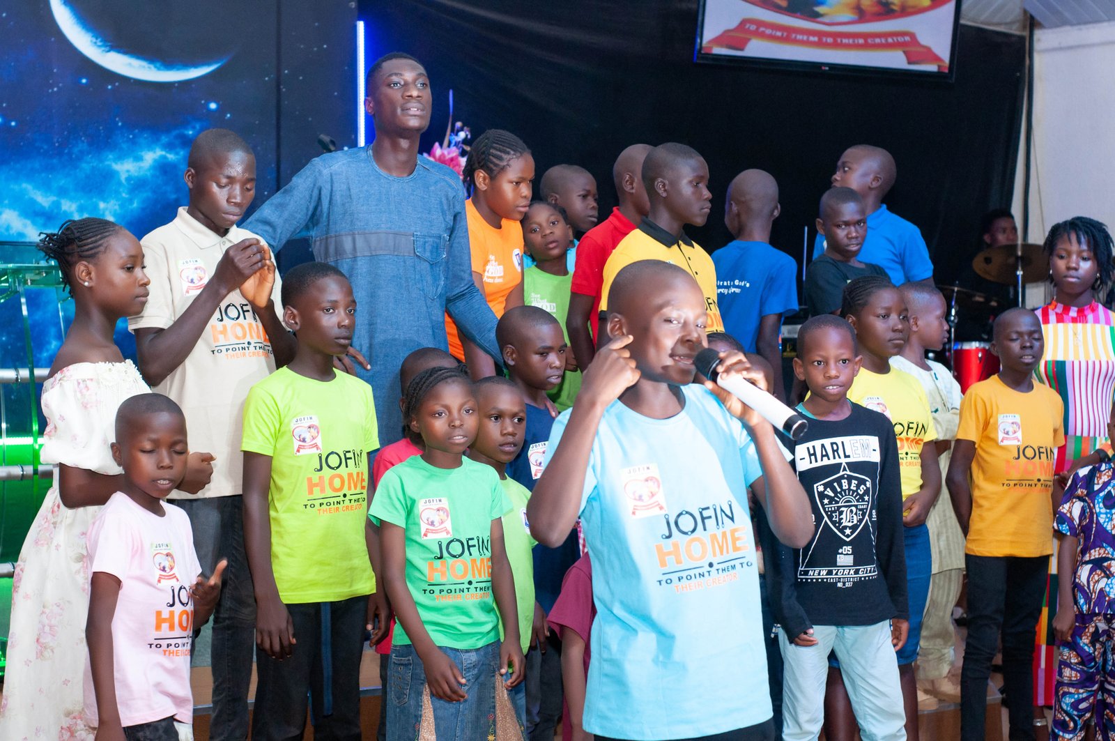 Children in JOFIN T-shirts on stage, one speaking into a microphone, at JOFIN's tenth-anniversary celebration in Jos