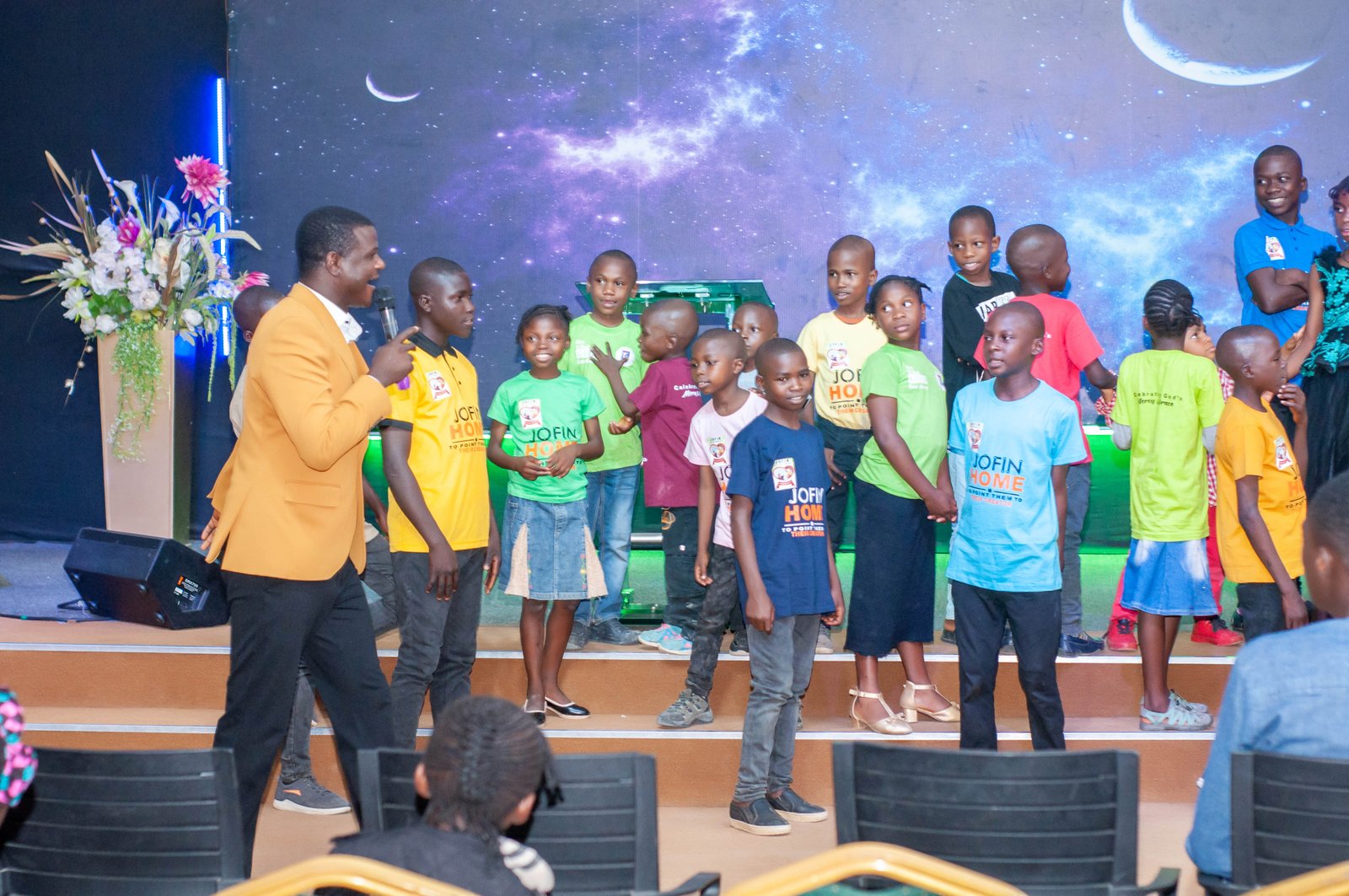 Children in JOFIN T-shirts standing on stage while a leader speaks